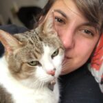 A woman presses her cheek gently against a calm tabby-and-white cat with green eyes, the two of them resting together in quiet connection.