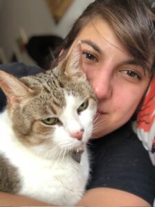 A woman presses her cheek gently against a calm tabby-and-white cat with green eyes, the two of them resting together in quiet connection.