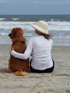 Cathy and her rescue dog Helen sitting together on beach, woman in white hat embracing golden retriever while watching ocean waves in who rescued who story from HARP Pittsburgh