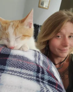 A soft orange-and-white cat naps peacefully on a plaid blanket, while a woman with a gentle smile sits beside him, close and content.