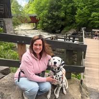 A woman kneeling outdoors beside a Dalmatian, both sitting near a wooden railing with trees and a covered bridge in the background.