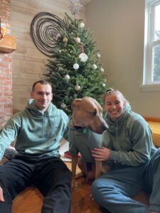 A couple sitting on the floor with their tan dog in matching green hoodies in front of a decorated Christmas tree.